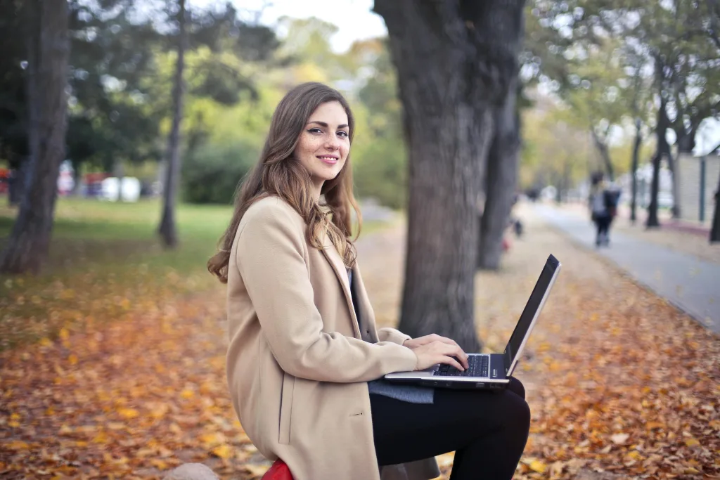 A young woman smiles while working on her laptop outdoors in an autumn park, surrounded by fallen leaves.